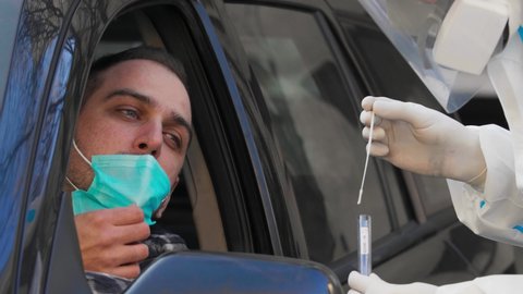 Medical worker performing drive-thru COVID-19 test, taking nasal swab sample from male patient through car window, PCR diagnostic for Coronavirus, doctor in PPE holding test kit.