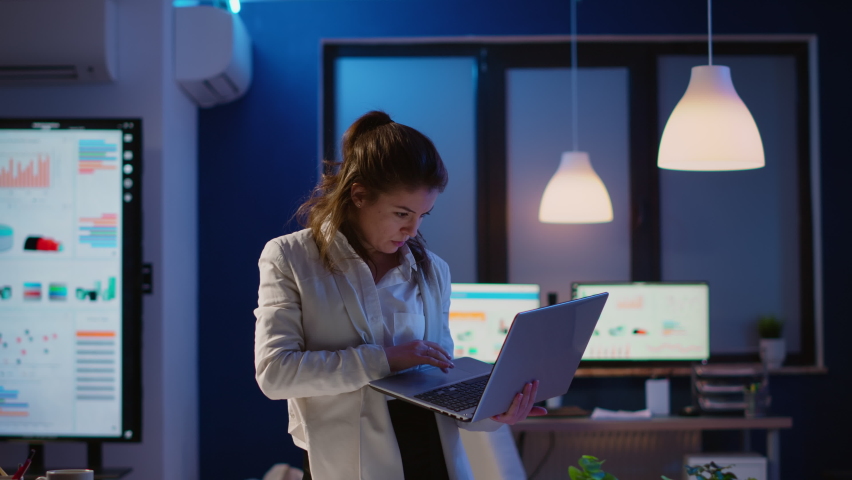 Focused manager woman checking business reports standing in startup office late at night holding laptop. Busy employee using technology network wireless doing overtime reading writing, searching | Shutterstock HD Video #1065410602