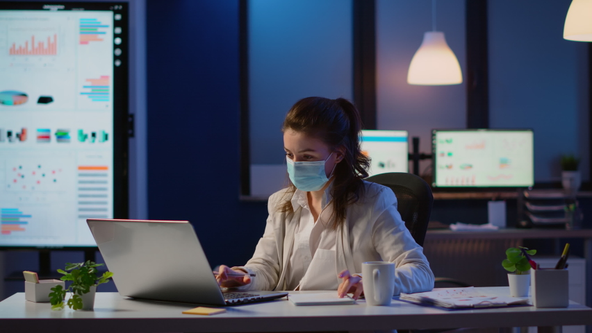 Manager woman with face mask working overtime in new normal business office checking team project, taking notes, analysing financial documents sitting at desk late at night during global pandemic | Shutterstock HD Video #1065410659