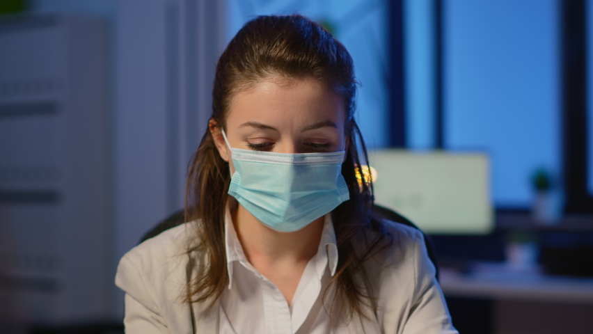 Close up of woman with face mask reading emails late at night to respect deadline of project working in new normal business office, taking notes, analysing documents overtime during global pandemic | Shutterstock HD Video #1065410812