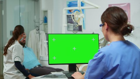Dentist nurse looking at green screen display while doctor exams patient lying on dental chair. Stomatologist assistant using monitor with green chroma key isolated display mock-up touchscreen