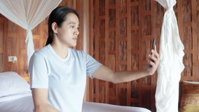 4K Asian smiling happy women use smartphone for video call online at hotel room. woman calling video chat at mobile phone in hotel. woman sitting on bed and making online video call at hotel villa