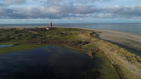 4K Aerial view slowly flying away from red lighthouse over water and dunes. People walking in the dunes of Dutch Island Texel. Birds eye landscape view with blue sky, big clouds and long shadows.