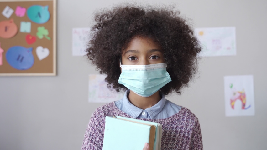 Cute small elementary reopen school pupil african american kid child girl wearing face mask looking at camera standing in classroom. Children safety for covid protection, headshot close up portrait. | Shutterstock HD Video #1065428290