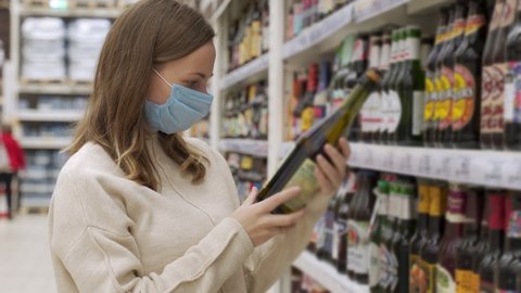 Female in a medical mask takes alcoholic drinks from the supermarket shelf. Shopping for alcohol in the store. The girl chooses a drink in the supermarket.