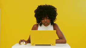 Young Afro-American woman is working at yellow laptop on yellow background, feeling under pressure because of big work loading, has some difficulties and gets nervous, creative video, Slow motion.