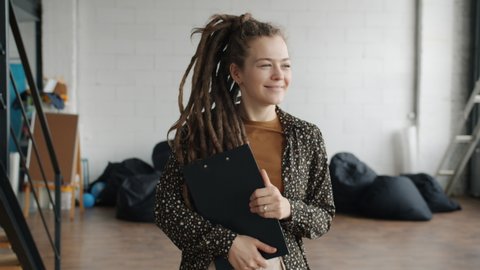 Dolly shot of attractive young woman with dreadlocks walking in office saying hi waving hand smiling. Coworking place and happy people concept.