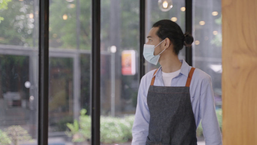 Young Asian man wearing protective face mask while working in restaurant or cafe and greeting his colleague by elbow touch. Alternative greeting for prevent spread of coronavirus infection. Royalty-Free Stock Footage #1065442954