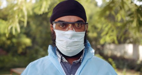 Slow-motion portrait of adult Sikh turbaned male medical staff wearing personal protective equipment gets ready for field hospital adjusts his face shield mask outdoors looking at camera pov.