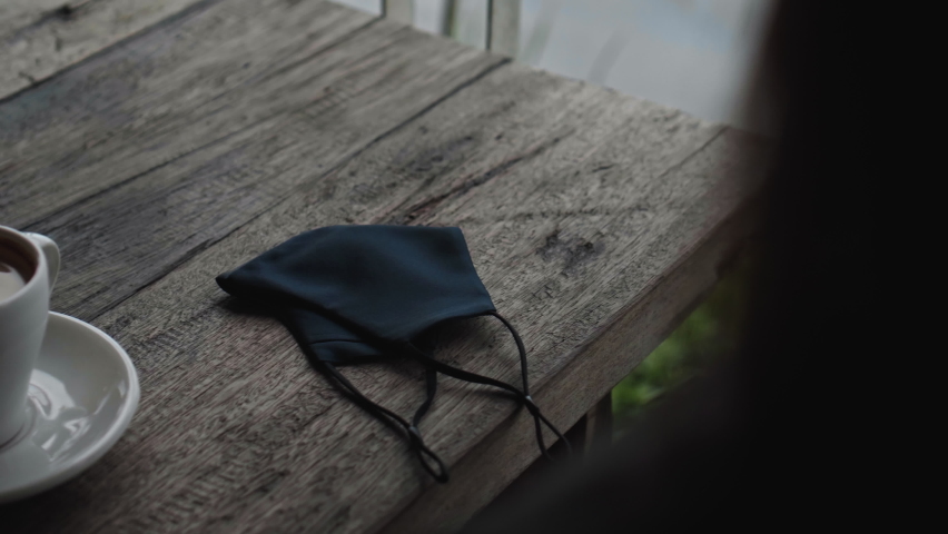 A young European girl sits at a table in a public place and takes a soft black protective mask from the table with her hands