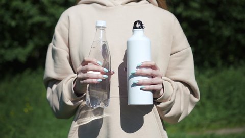 Young beautiful woman is showing water bottles while standing in city park on summer spbd. Front view of caucasian female holding plastic products in her hands, supporting idea of environmentally