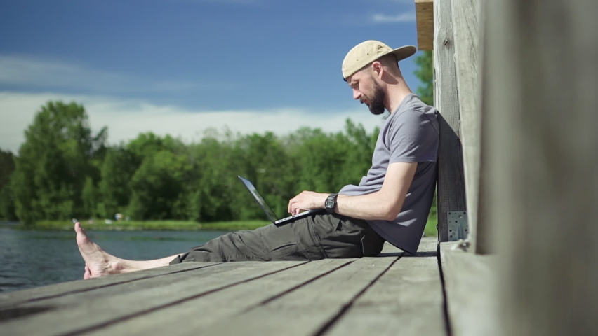 guy with laptop sit on lake pier and types on computer Spbd. man outside works remotely. carefree man resting and uses laptop on pond. concept education, student Royalty-Free Stock Footage #1065446128