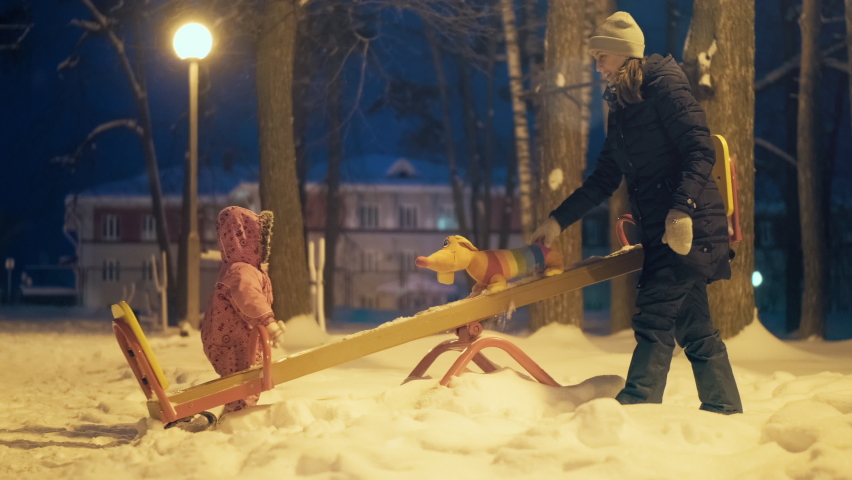 Kid on the playground. Toddler girl has fun on the swing on the winter playground at night with her mother