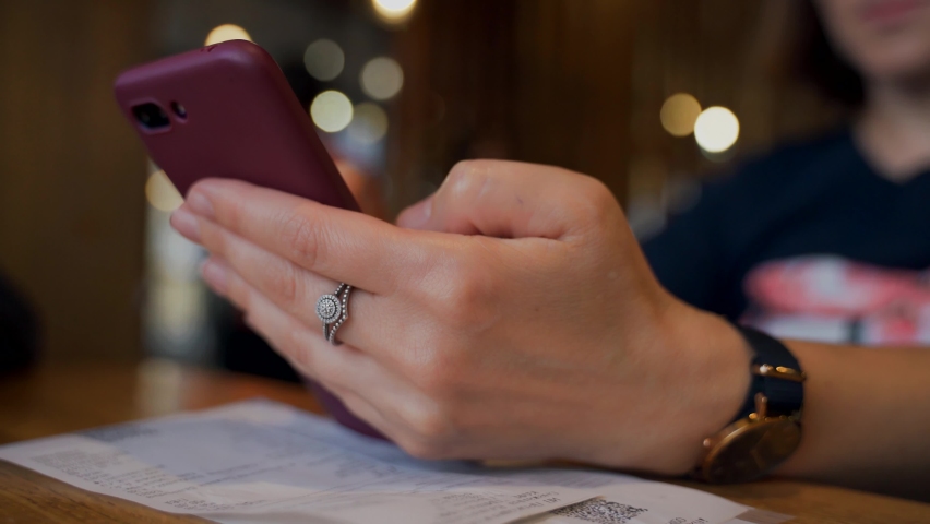 A girl with a phone in her hands sits at a table in a cafe while waiting for an order. Woman's hand with a smartphone close-up.