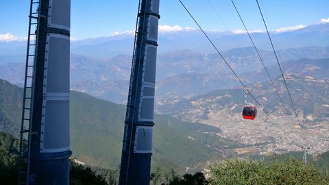 Red colored cable car moving up the hill near Chandragiri, Nepal with stunning panoramic view over Kathmandu valley and the snow-capped peaks of the Himalayas on sunny day.
