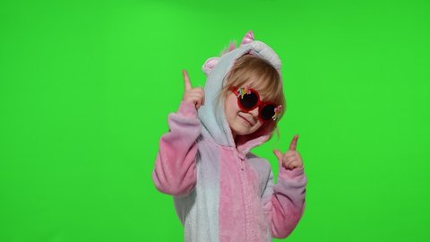 Portrait of a cute young little child in sunglasses smiling, dancing in unicorn costume on chroma key green background. Kid girl animator making gun gesture with hands in unicorn pajamas. Slow motion