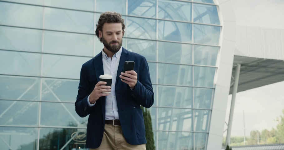 Smart casual dressed Office Worker is drinking Coffe and using his Phone. Man is standing near modern office Building and browsing Social Networks at the Break. Coffe Break. Office Style. Startup. Royalty-Free Stock Footage #1065457633