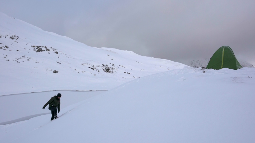 A man walking up a snowy mountain to his wild camping tent on the snow