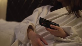 Woman white bathrobe lies soft bed her room, resting hotel room after trip. Watching SmartTV, popular blogger program. Switches channels, makes video volume higher, lower. Cozy relaxed time before bed