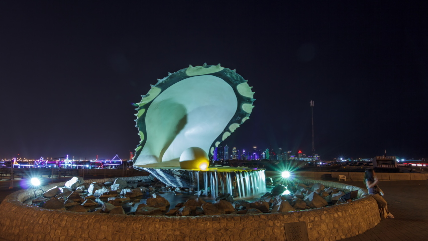 Corniche with fountain featuring an oyster with a gigantic pearl inside illuminated by night timelapse hyperlapse with the Doha skyline and boats behind it. People walking around Royalty-Free Stock Footage #1065464053