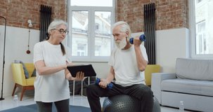 Adorable positive active mature couple in sportswear doing exercises with dumbbells simultaneously watching online lessons on tablet device