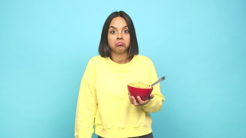 Young latin woman eating a cereals bowl shrugs shoulders and open eyes confused