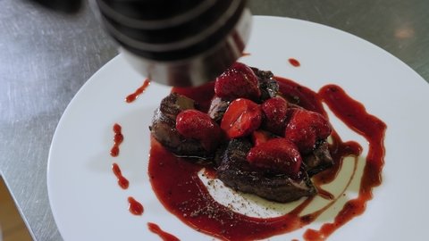 Close-up of the chef serving dishes in the restaurant kitchen, pouring juicy beef steak sauce with strawberries and sprinkle with ground pepper.