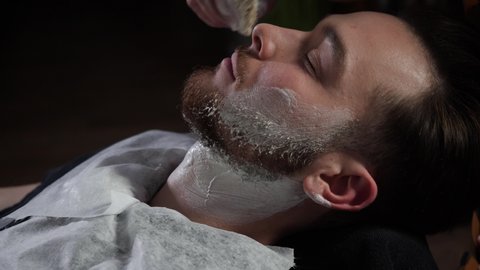 Barber works with shaving brush. Close-up of man getting his beard lathered with shaving cream in barbershop. White foam on client's face before shave Beard styling. High quality 4k footage