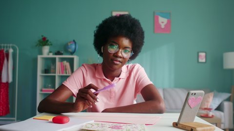 Portrait of young woman, stylish student in pink T-shirt and cool glasses, sitting at desk at home and playing with purple pencil, looking at camera and smiling, Zoom out, Slow motion.