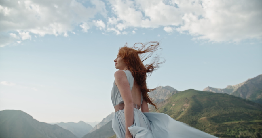 Gorgeous caucasian girl wearing a light dress is posing for a photo shoot on scenic mountain background while wind is blowing her red hair 4k footage Royalty-Free Stock Footage #1065486481