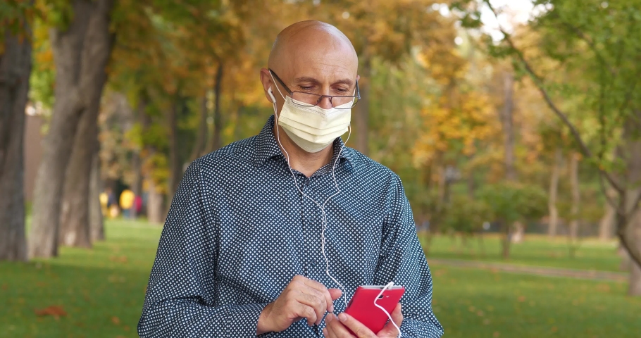 Man in medical protective mask and glasses sits on the bench in public garden and communicates via smartphone by video link. Cinema 4K 60fps video