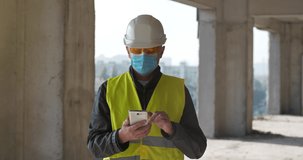 Builder in medical protective mask and hard hat stands at construction site and communicates via smartphone by video link, shows to interlocutor the construction site. Cinema 4K 60fps panoramic video