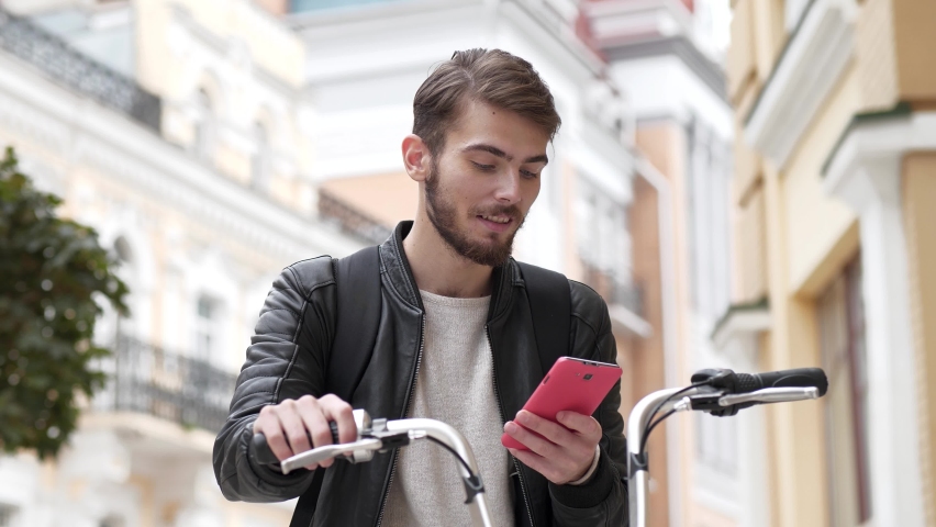 Slow Motion Guy in black leather jacket stands on street with bicycle and reads a message on his smartphone, then he celebrates victory and shows violent emotions, he became a winner
