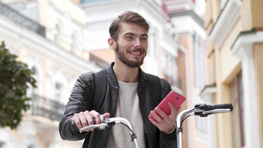 Slow Motion Guy in black leather jacket stands on street with bicycle and reads a message on his smartphone, then he celebrates victory and shows violent emotions, he became a winner
