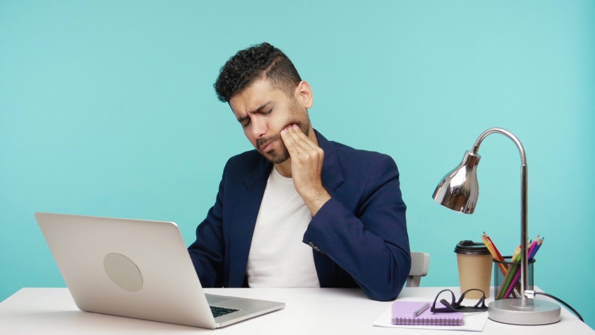 Upset depressed office worker touching cheek working on laptop, feeling acute tooth pain, problems with teeth and gums. Indoor studio shot isolated on blue background | Shutterstock HD Video #1065490033