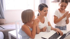 Cheerful woman with two kids sitting at table in living room and using computer