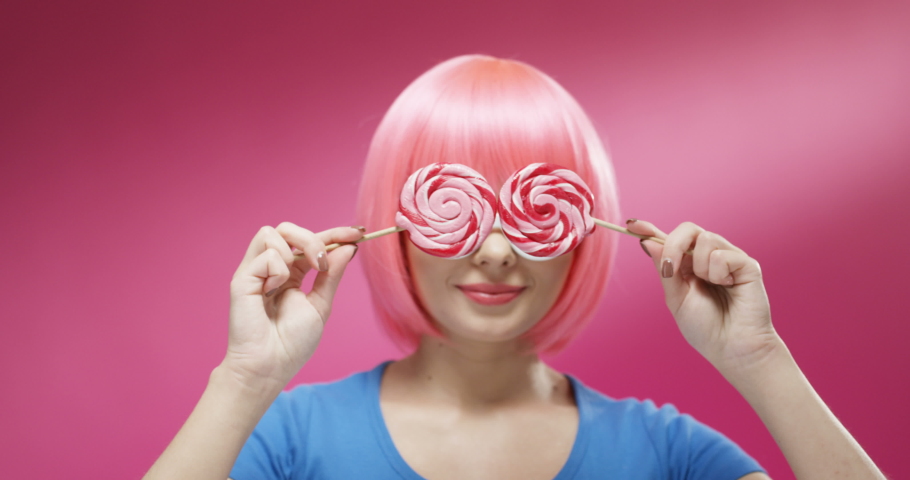 Close up of pretty Caucasian young woman in stylish sunglasses closing eyes with two lollypops and smiling cheerfully. Portrait of beautiful happy girl playing with candies on bright wall background.