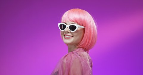 Стоковое видео: Portrait of Caucasian joyful stylish woman in pink wig and fancy sunglasses turning face and smiling to camera on velvet background. Close up of happy girl with smile. Extraordinary Barbie female.