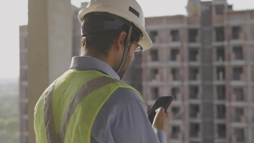 slow motion close up shot of a young male Asian civil engineer wearing hard and safety jacket standing on a top of a building near a construction site using mobile phone to type a text message  Royalty-Free Stock Footage #1065514051