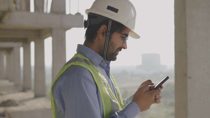 Slow motion close up shot of a young male Asian civil engineer wearing hard and safety jacket standing on a top of a building near a construction site using mobile phone to type a text message  | Shutterstock HD Video #1065514051