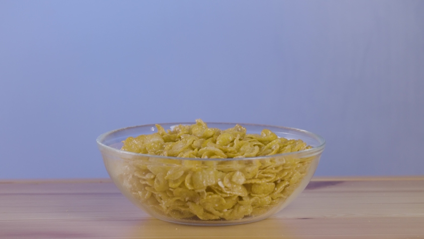 Pouring milk over a transparent bowl full of cornflakes with drops of milk falling on a table. Action. Tasty breakfast with cereal.