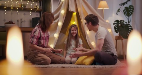 Happy young family sitting on floor with teepee and embracing. Portrait of cheerful mother, father and daughter hugging and relaxing on carpet near wigwam spending time together at home