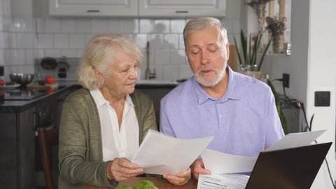 A sixty-year-old wife and husband sit at a desk and laptop, holding bills in their hands, managing the family budget, checking utility bills.