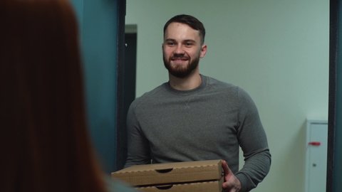 Smiling bearded delivery man delivering boxes with hot pizza to redhead woman customer on doorstep at apartment. Back view of female receiving parcel at home from courier male at front door.
