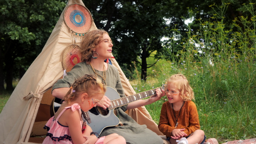 Boho family. Mom plays the guitar with her two daughters in nature. Hippie family is having fun around the wigwam.