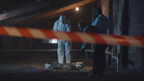 Two policemen and forensic specialist taking photos of evidence and corpse on crime scene. Detectives and criminologist in safety overall examining body behind caution tape