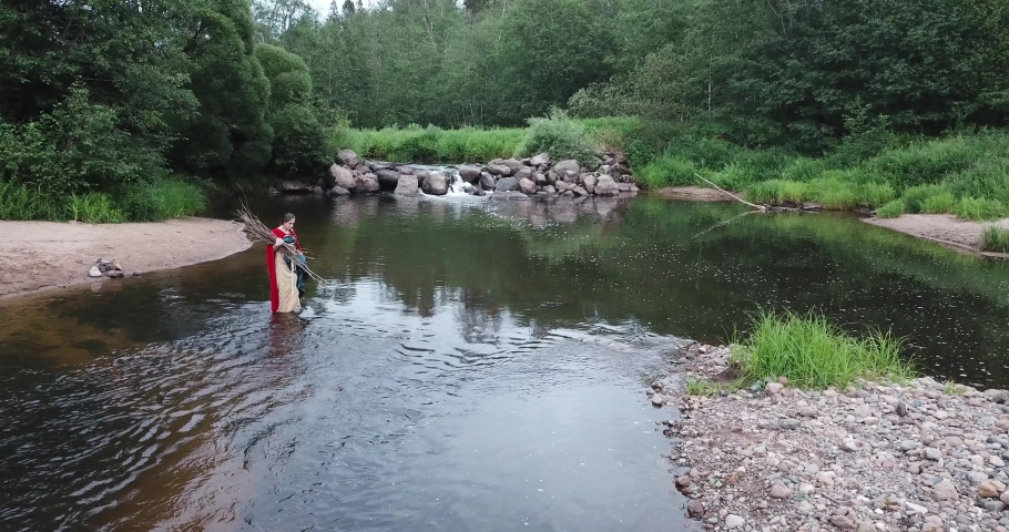 girl in red cloak crosses river into ford, carries sticks, branches, firewood in her hands. Walking over stones along river bank with a waterfall. Forest, evening. Fabulous image, fairytale character.