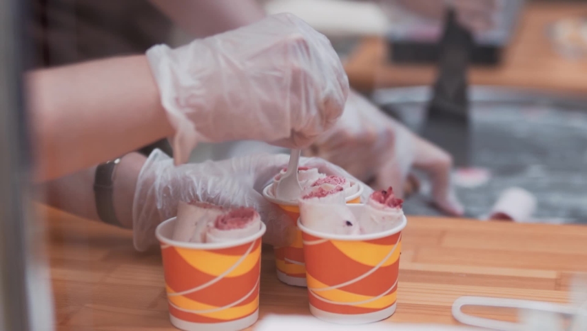 Thai fried ice cream into rolls in a colored cup decorate with marshmallows hands in gloves with food tongs