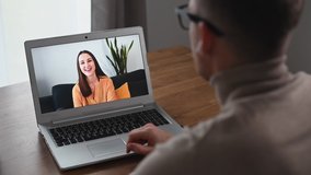 A young cheerful woman is waving hello on the laptop screen, a guy is greeting too. Video call with friend, dating online. Close-up above shoulder view