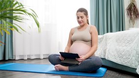 Young pregnant woman sitting on fitness mat and practicing yoga online on tablet computer. Concept of healthy lifestyle, healthcare and sports during pregnancy.